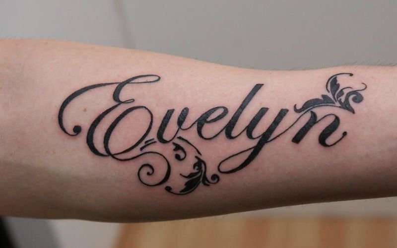 A tattoo on a forearm features the name Evelyn in elegant, cursive script with decorative flourishes and swirls around the letters.