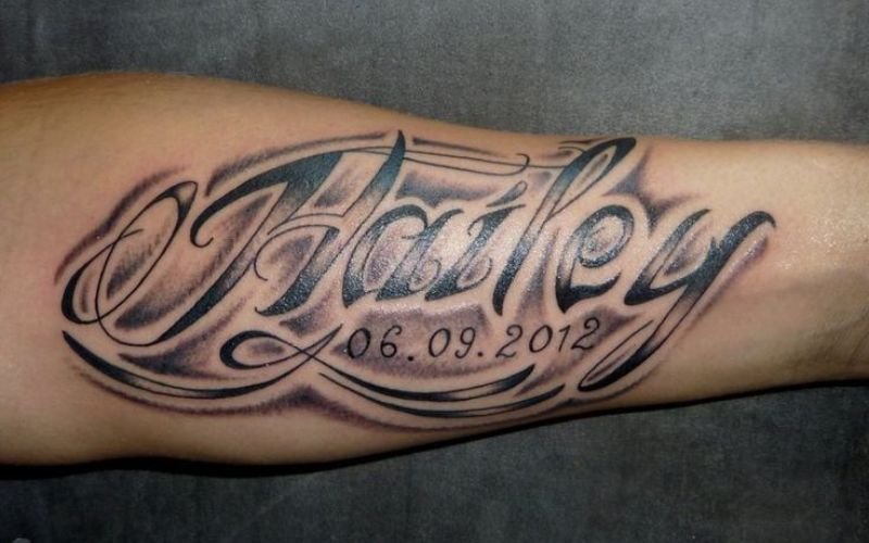 A forearm tattoo features the name Hailey in large, decorative script with the date 06.09.2012 written underneath, all surrounded by shaded, smoky effects.